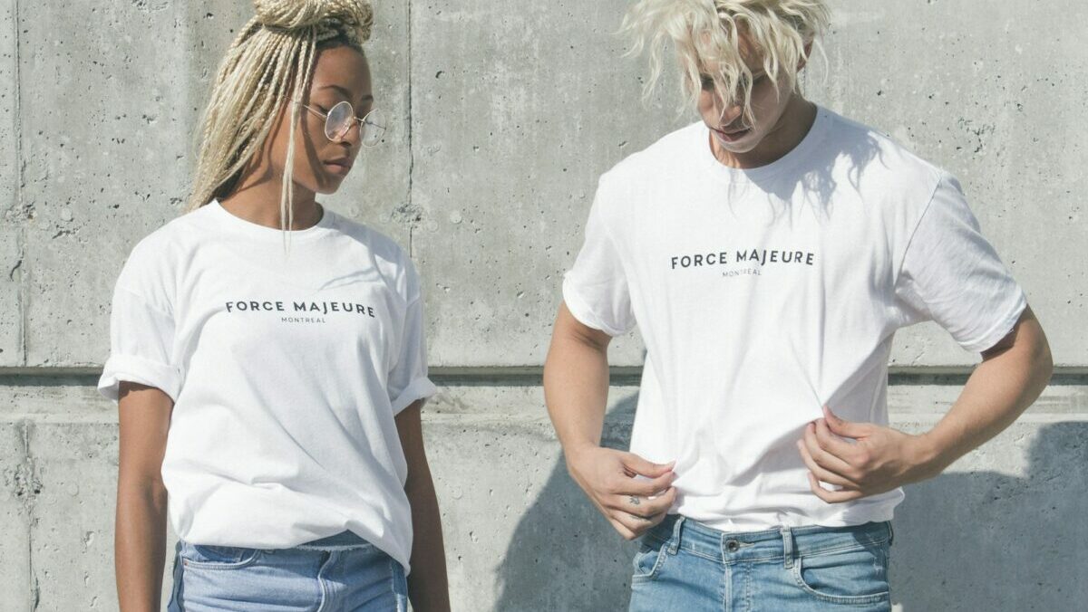 man and woman in white-and-gray force majeure-printed crew-neck t-shirts standing near gray concrete wall